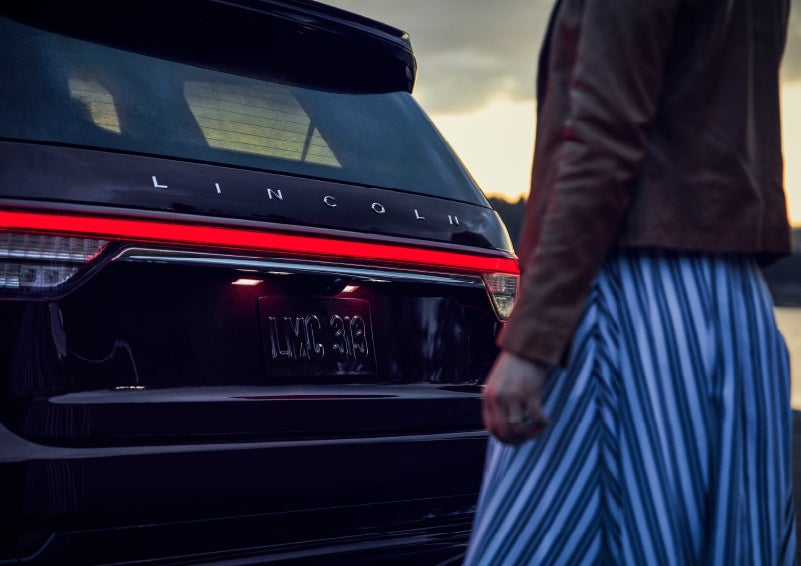 A person is shown near the rear of a 2024 Lincoln Aviator® SUV as the Lincoln Embrace illuminates the rear lights | Parkway Lincoln in Dover OH