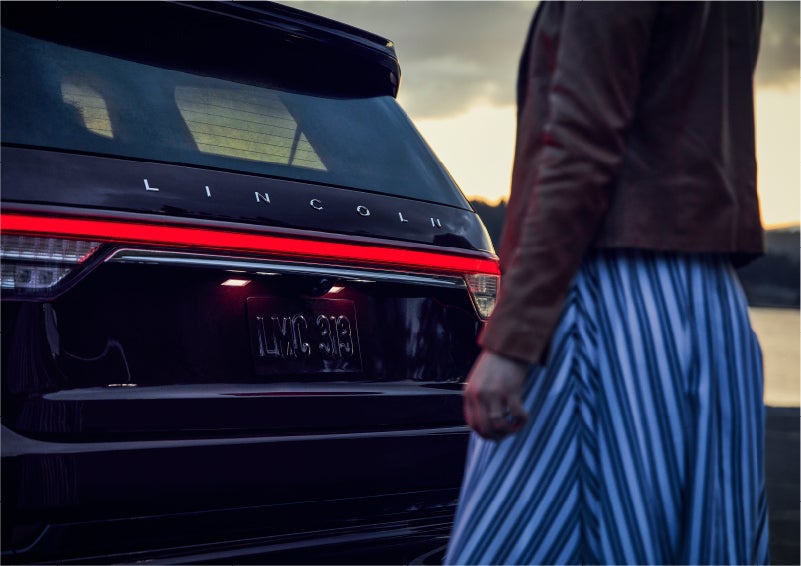 A person is shown near the rear of a 2023 Lincoln Aviator® SUV as the Lincoln Embrace illuminates the rear lights | Parkway Lincoln in Dover OH
